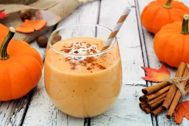 A creamy Spiced Pumpkin Coffee smoothie in a rounded glass, topped with whipped cream and cinnamon, with a striped straw. Surrounded by mini pumpkins, cinnamon sticks, and autumn leaves on a rustic wooden surface, evoking a cozy fall vibe.