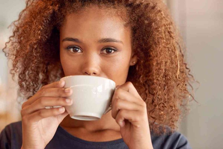 A woman with curly hair holds a white mug with both hands and looks directly at the camera while taking a sip.