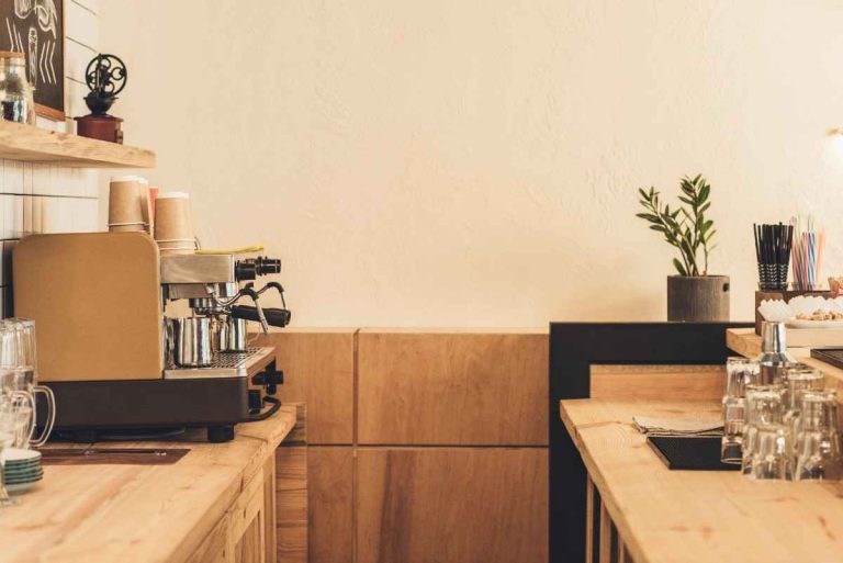 A cozy coffee shop counter with a coffee machine, stacked cups, glass mugs, a potted plant, and colorful straws on a wooden surface against a light-colored wall.