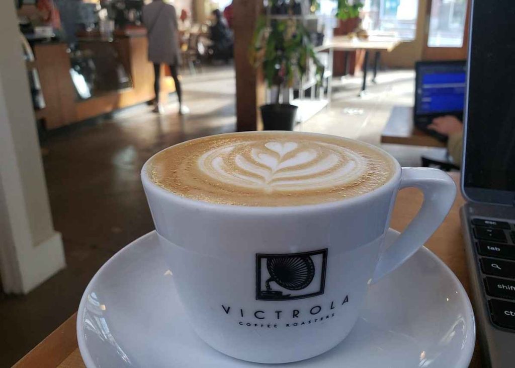 A cup of latte with heart-shaped foam art sits on a saucer at a cafรฉ table, near a laptop. The cafรฉ interior is bright, with patrons in the background and sunlight streaming through windows.